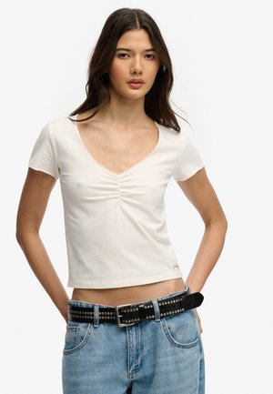 Kobieta z długimi ciemnymi włosami, ubrana w biały marszczony top z krótkim rękawem, włożony w niebieskie dżinsy z czarnym paskiem z nitami.