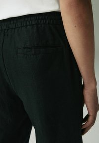 Pantaloni negri din in, cu un elastic la talie și un singur buzunar la spate. Textura pare moale și respirabilă.