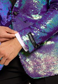 Blazer viola con paillettes e dettagli iridescenti, bottoni neri, polsini bianchi e un'etichetta con la scritta "OPPOSUITS". Superficie testurizzata che riflette la luce.