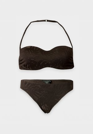 Kvinne står barbeint iført en mørkebrun to-delt bikini med stroppeløs topp og smale underdeler mot en ensfarget hvit bakgrunn.