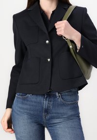 Granatowa krótka blazerka z dwoma dużymi przednimi kieszeniami, srebrnymi guzikami i dopasowanym krojem, zestawiona z niebieskimi dżinsami i zieloną torbą.