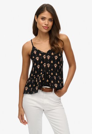 Mujer con una blusa negra sin mangas con estampado floral y botones, y pantalones blancos, de pie con una mano en el bolsillo contra un fondo liso.