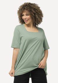 Maglietta corta verde chiaro con scollo quadrato, vestibilità rilassata e texture morbida, indossata con pantaloni neri.