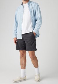 Vaaleansininen napillinen paita valkoisen t-paidan päällä, paritettu tummien shortsien ja beigejen lenkkareiden kanssa. Ominaisuuksia ovat puuvillamateriaali, rento muotoilu ja sekoitetut tekstit.