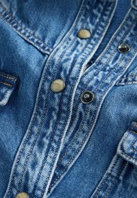 Jeansskjorte med metal tryknapper, blå stof, og synlige syningsdetaljer. Har lommer og en struktureret finish.