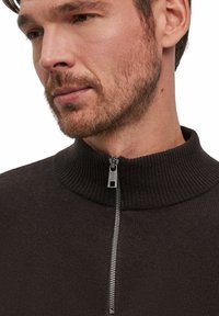 Polo a maglia marrone scuro con un alto colletto a coste e zip argentata. Il tessuto sembra morbido con una superficie liscia. Vista ravvicinata della parte superiore del torso.