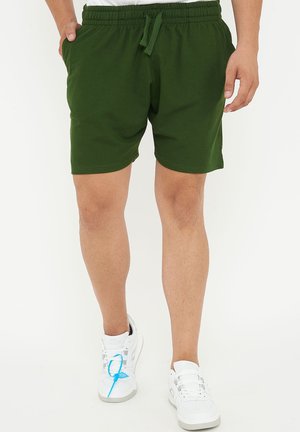 Mann trägt dunkelgrüne Shorts mit Kordelzug und weiße Sneakers, steht mit einer Hand in der Tasche vor weißem Hintergrund.
