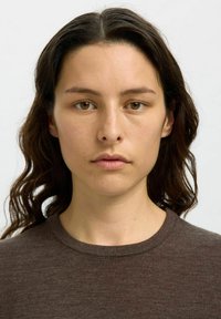 Donna con capelli scuri, ondulati e di lunghezza alle spalle, che indossa un maglione a collo tondo di colore marrone con una texture. Pelle chiara, occhi marroni, espressione neutra.