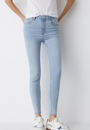 Person iført lyseblå skinny jeans, hvide sneakers og en hvid indstukket t-shirt, stående med hænderne i lommerne.