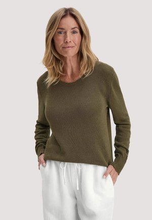 Vrouw met schouderlang blond haar, gekleed in een olijfgroene trui en witte broek met trekkoord, handen in de zakken, neutrale achtergrond.