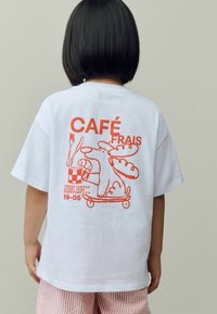 Vit bomull t-shirt med en orange cartoon-grafik av en hund med mat och en kaffekopp, texten "CAFÉ FRAIS" ovanför. Kortärmad.