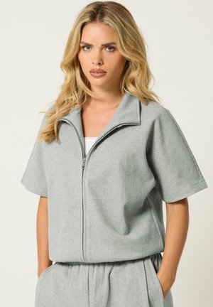 Femme aux longs cheveux blonds portant un haut gris clair zippé à manches courtes et un pantalon assorti, debout avec une main dans la poche.