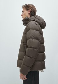 Hnědá prošívaná bunda s kapucí, ozdobená detaily z pružného šňůrky, prošívaným vzorem a patentními knoflíky. Boční pohled ukazuje hranatý střih.