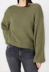 Olivengrön stickad tröja med bred halsringning, lösa ärmar och ribbat avslut; parat med mörkgrå tajta jeans.