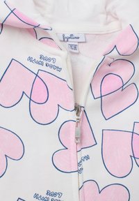 Sudadera blanca con cremallera y patrones de corazones rosas contorneados en azul. La tela es suave; cuenta con una cremallera plateada y una etiqueta cosida en el interior.