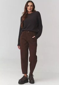 Kobieta z długimi brązowymi włosami nosi czarny sweter, brązowe spodnie z wysokim stanem i czarne buty na platformie, stojąc z jedną ręką w kieszeni.