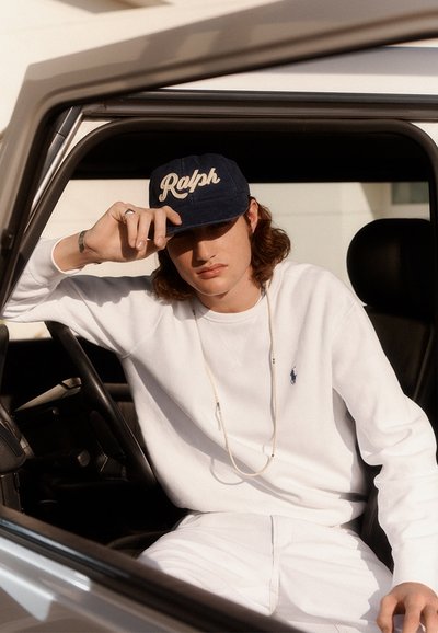 Jonge man die een witte trui en een donkerblauwe pet met 'Ralph' draagt, zit in een auto, houdt de klep van de pet vast en kijkt naar beneden.