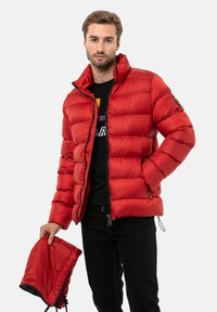 Rote Daunenjacke mit durchgehendem Reißverschluss, hohem Kragen und elastischen Bündchen. Inklusive abnehmbarer Kapuze. Das Model hält eine passende Kapuze.