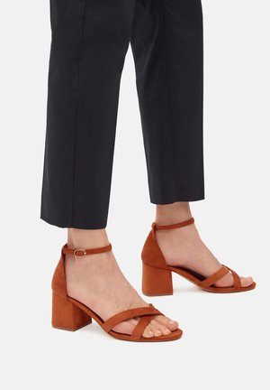 Bein einer Frau, die rostorange Wildleder-Sandaletten mit Blockabsatz und Knöchelriemen sowie schwarze gekürzte Hosen auf hellem Hintergrund trägt.