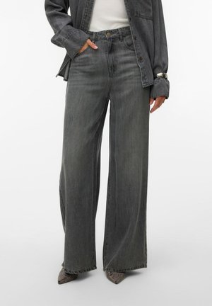 Persona che indossa jeans grigi a vita alta e a gamba larga, giacca di jeans grigia sopra un top bianco, con una mano in tasca e scarpe con punte visibili.