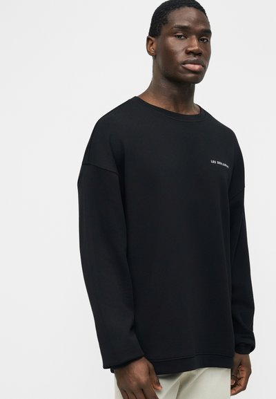 Νέος άνδρας που φοράει ένα χαλαρό μαύρο πουκάμισο με μακριά μανίκια και λευκό κείμενο "LES BENJAMINS" στο στήθος, στέκεται μπροστά σε μια απλή ανοιχτή επιφάνεια.