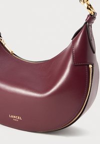 Sac à épaule en cuir bordeaux profond avec des accents de fermeture éclair dorés et un fermoir en métal doré, arborant le logo Lancel Paris à l'avant.