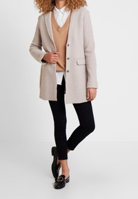 Donna con cappotto beige sopra un maglione color sabbia e camicia bianca, pantaloni skinny neri e mocassini neri con fibbia dorata, in piedi davanti a uno sfondo bianco.
