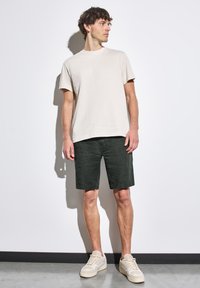 Světle béžové tričko s krátkým rukávem v kombinaci s tmavě zelenými kraťasy. Model má na sobě béžové tenisky. Jednoduchý, ležérní a volný střih.