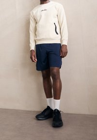 Krémová mikina s malým logem, černým zipem, námořnické šortky, bílé ponožky a černé sportovní boty. Minimalistický design, pohodlný střih.