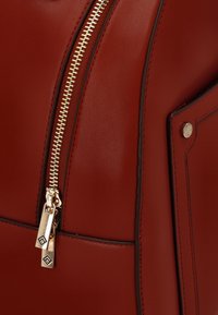 Sac en cuir rouge avec une fermeture éclair dorée, des surpiqûres décoratives et une poche latérale avec un détail de bouton-pression en métal. Texture lisse sur l'ensemble.