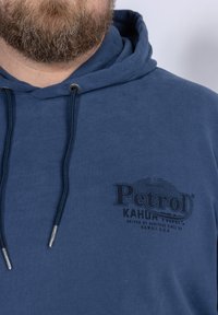 Felpa blu navy realizzata in tessuto morbido con una grafica frontale che riporta la scritta "PetroD KAHUA." Presenta un cappuccio con lacci e punte in tono argento.