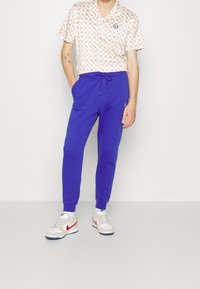 Avslappnad outfit med en mönstrad kortärmad skjorta i kräm och röd, tillsammans med blå byxor med dragsko och vita sneakers.