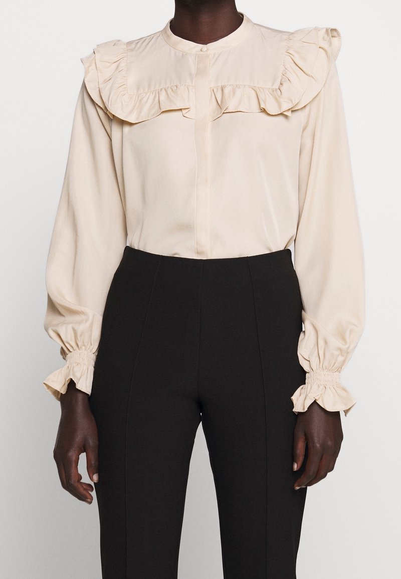 Blouse beige clair avec col et poignets volantés, dotée d'une fermeture à boutons. Manches longues et tissu lisse, associée à un pantalon noir taille haute.