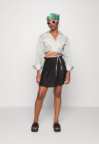 Camisa cinza abotoada cropped com dois bolsos na frente, saia preta plissada com cintura ajustável por cordão e sandálias slide pretas. Lenço de cabeça estampado em verde.