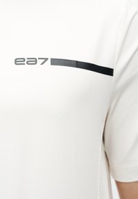 Άσπρο πουκάμισο με μαύρο λογότυπο "ea7" και οριζόντια μαύρη γραμμή κατά μήκος του πάνω μέρους του στήθους κοντά στον ώμο στη δεξιά πλευρά.