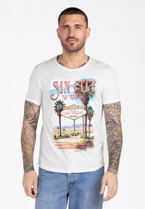 Uomo con capelli corti e tatuaggi sulle braccia, indossa una t-shirt bianca con grafica "Sin City Las Vegas" e jeans blu, in piedi davanti a uno sfondo bianco.