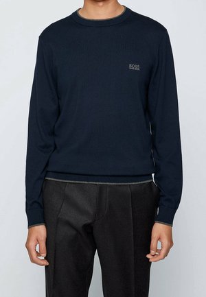 Mężczyzna ubrany w granatowy sweter z długim rękawem i okrągłym dekoltem z logo "BOSS HUGO BOSS" na klatce piersiowej oraz ciemnoszare spodnie.