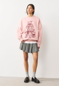 Ružová mikina s grafikou s ikonou naskladaných koktailových pohárov s mašličkami, v kombinácii s plisovanou čierno-bielou kockovanou sukňou a sivými ponožkami.