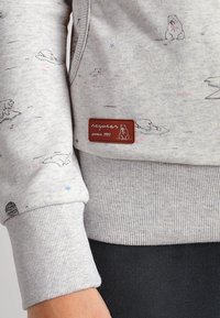 Harmaa collegepaita, jossa on jääkarhukuosiset, ribbaudet hihat ja ruskea nahkalappu, jossa lukee "raquwear vuodesta 1997". Paitaa elävöittävät hajallaan olevat siniset yksityiskohdat.