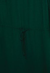 Tecido verde com uma textura suave, apresentando uma cintura ajustada amarrada com um cordão à condizer. Design simples sem padrões adicionais.