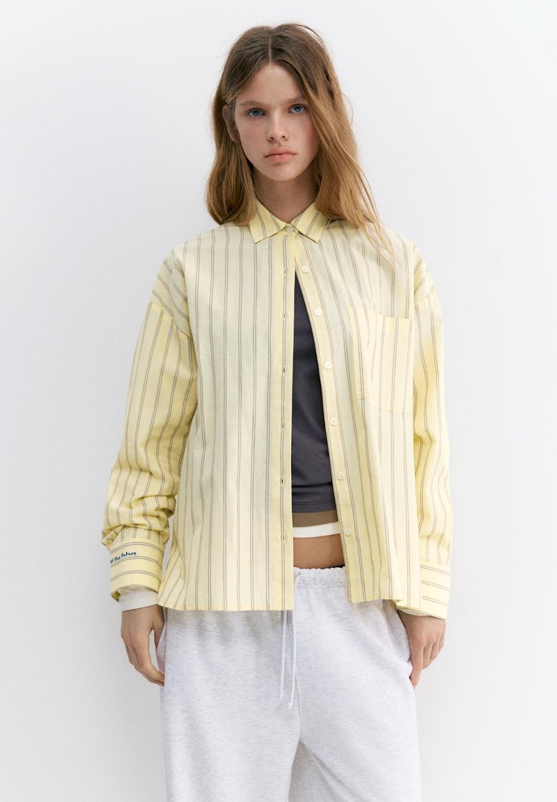 Jeune femme aux longs cheveux porte une chemise ample jaune rayée à boutons par-dessus un top sombre et un pantalon blanc à cordon, sur un fond uni.
