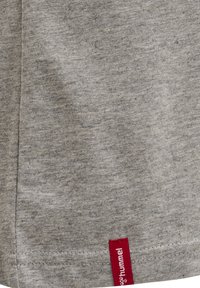 Gros plan d'un tissu gris avec une petite étiquette rouge portant la mention "hummel" cousue près de l'ourlet surpiqué.