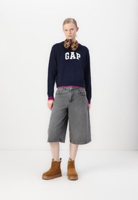 Pulover marin cu logo "GAP", șorturi de denim gri și cizme de gleznă din sueded maro cu tălpi groase. Accentele includ o margine roz la tivul puloverului.