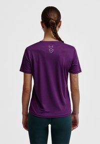 Lila kortärmad tröja med rund halsringning och slät textur. Har tre vita nedåtvända pilar på övre delen av ryggen.