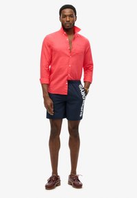 Chemise en lin rouge à boutons, shorts de bain bleu marine avec logo blanc, et chaussures en cuir bordeaux. Les shorts ont des poches latérales et une taille élastique avec cordon.