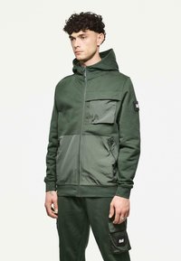 Groene zip-up hoodie met een gestructureerde stof, voorzien van een grote voorkantzak, contrasterende panelen en een verstelbare capuchon.