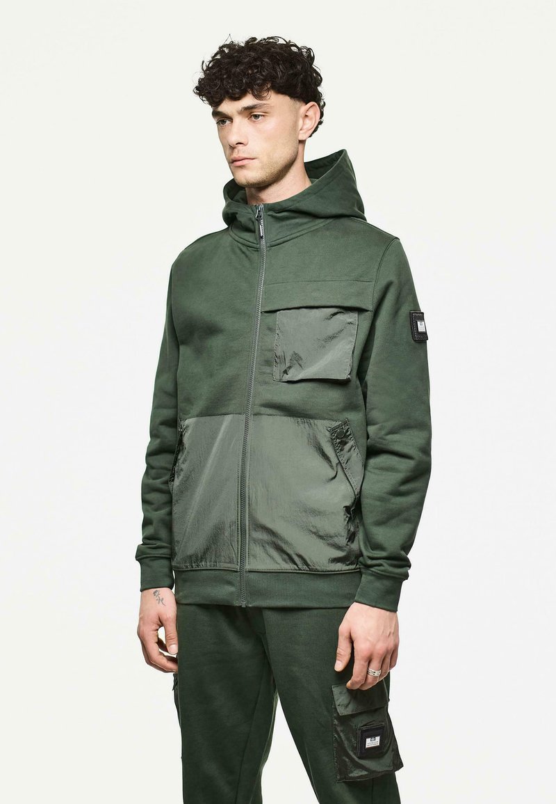 Groene zip-up hoodie met een gestructureerde stof, voorzien van een grote voorkantzak, contrasterende panelen en een verstelbare capuchon.