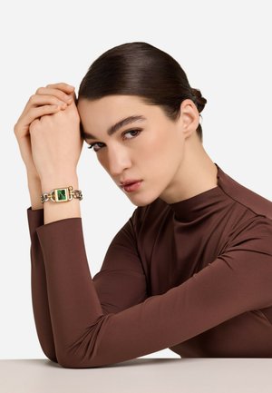 Zlati uhani z zelenim pravokotnim kamnom, debelimi verižnimi povezavami, nosijo na zapestju. Vidna je tudi modelova rjava oprijeta majica z dolgimi rokavi.