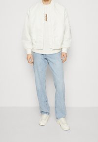 Uomo che indossa una giacca bomber bianca, una felpa con logo beige chiaro, jeans blu chiaro e sneakers bianche, in piedi su uno sfondo bianco.