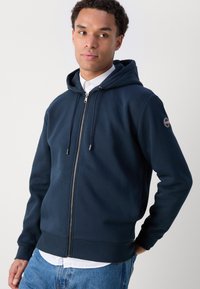 Felpa con zip di colore blu navy in tessuto morbido, con cappuccio regolabile tramite cordino, polsini e orlo a coste, caratterizzata da un patch con logo sulla manica sinistra.